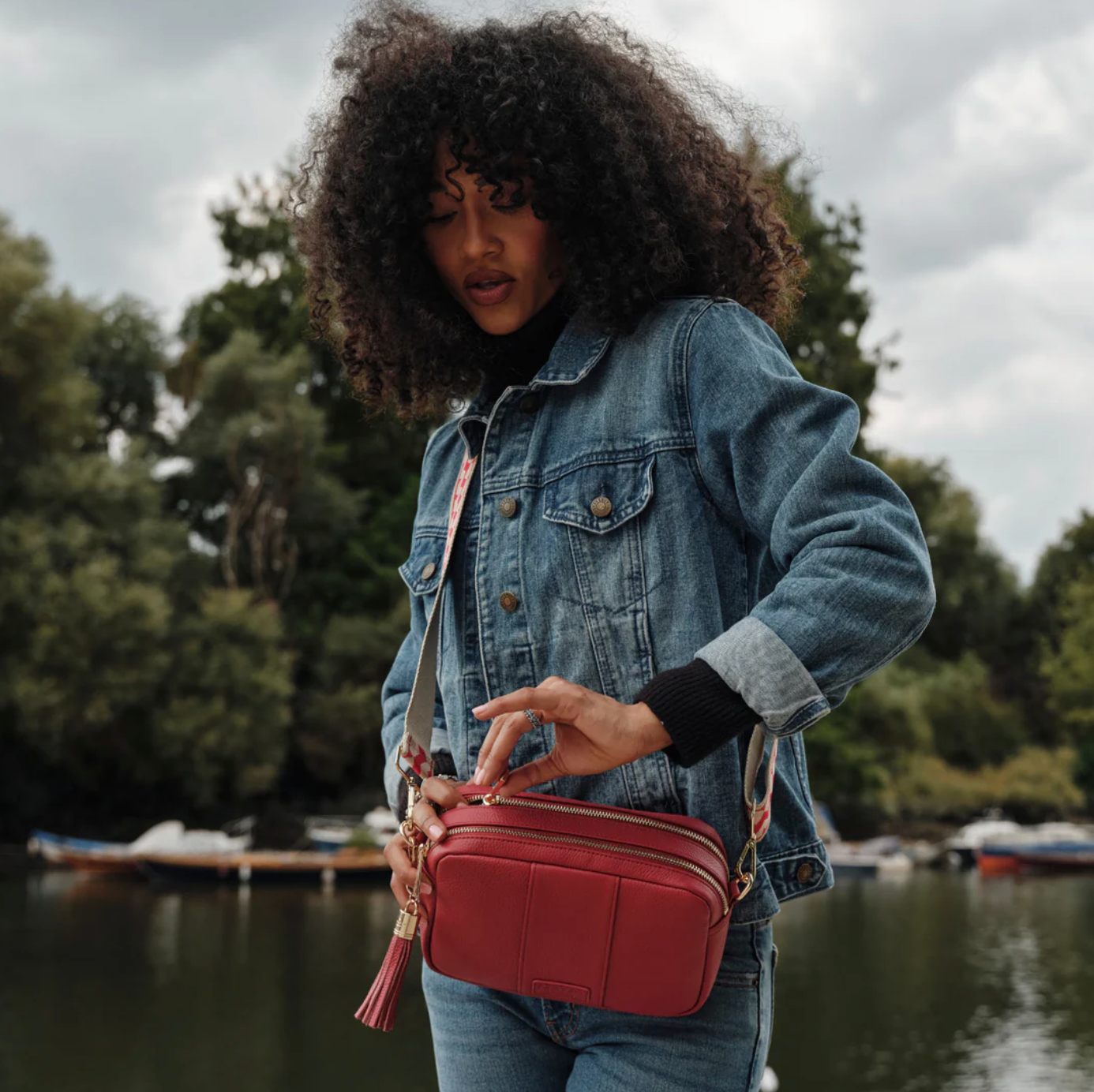 Woman holding a red handbag by a body of water with trees in the background