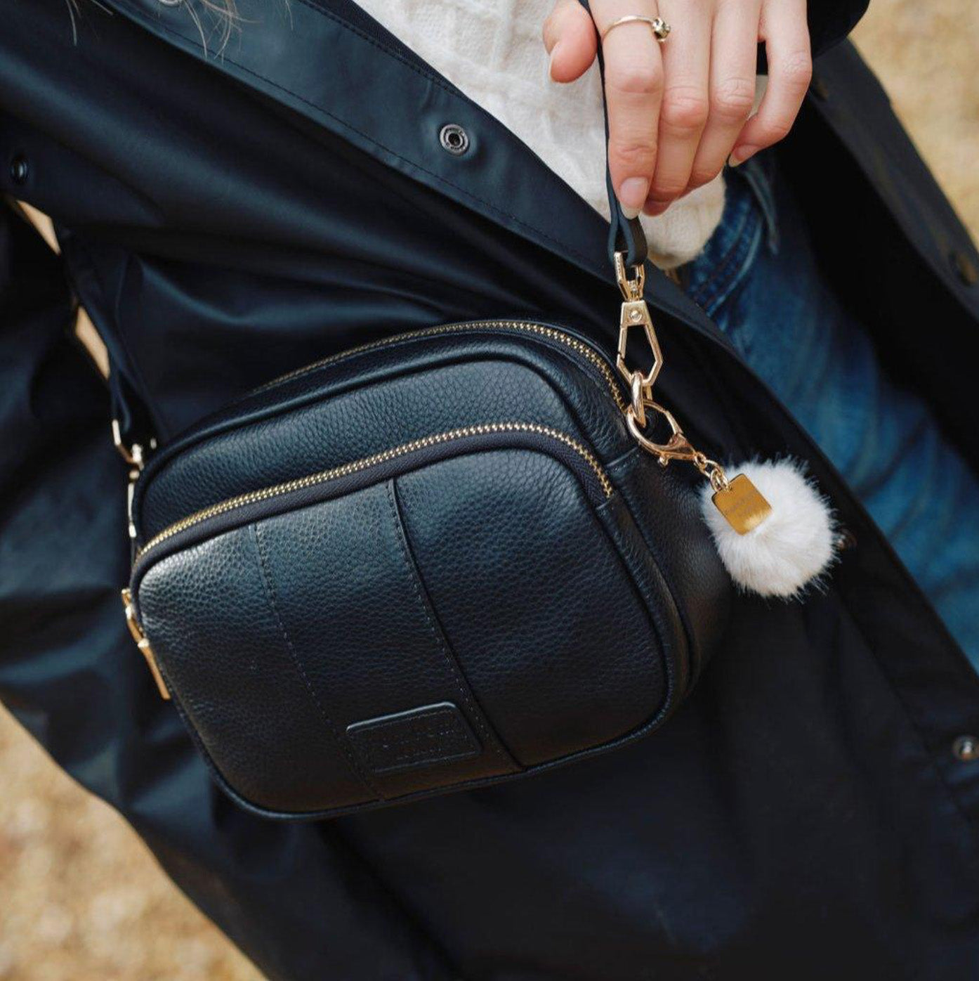 Black leather bag with a gold chain and white pom-pom charm held by a person wearing a dark jacket.