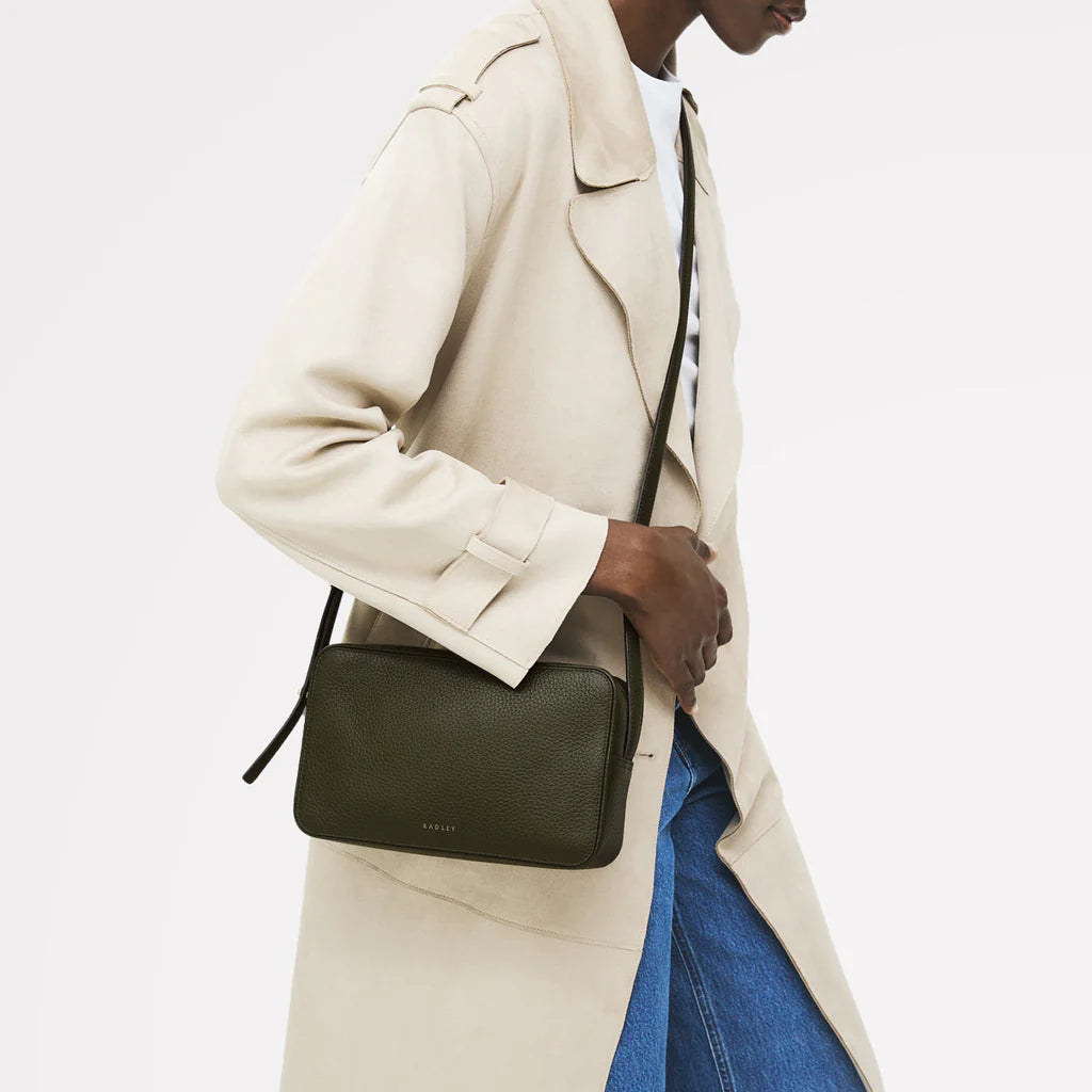 Person wearing a beige coat and carrying a green crossbody bag on a light gray background