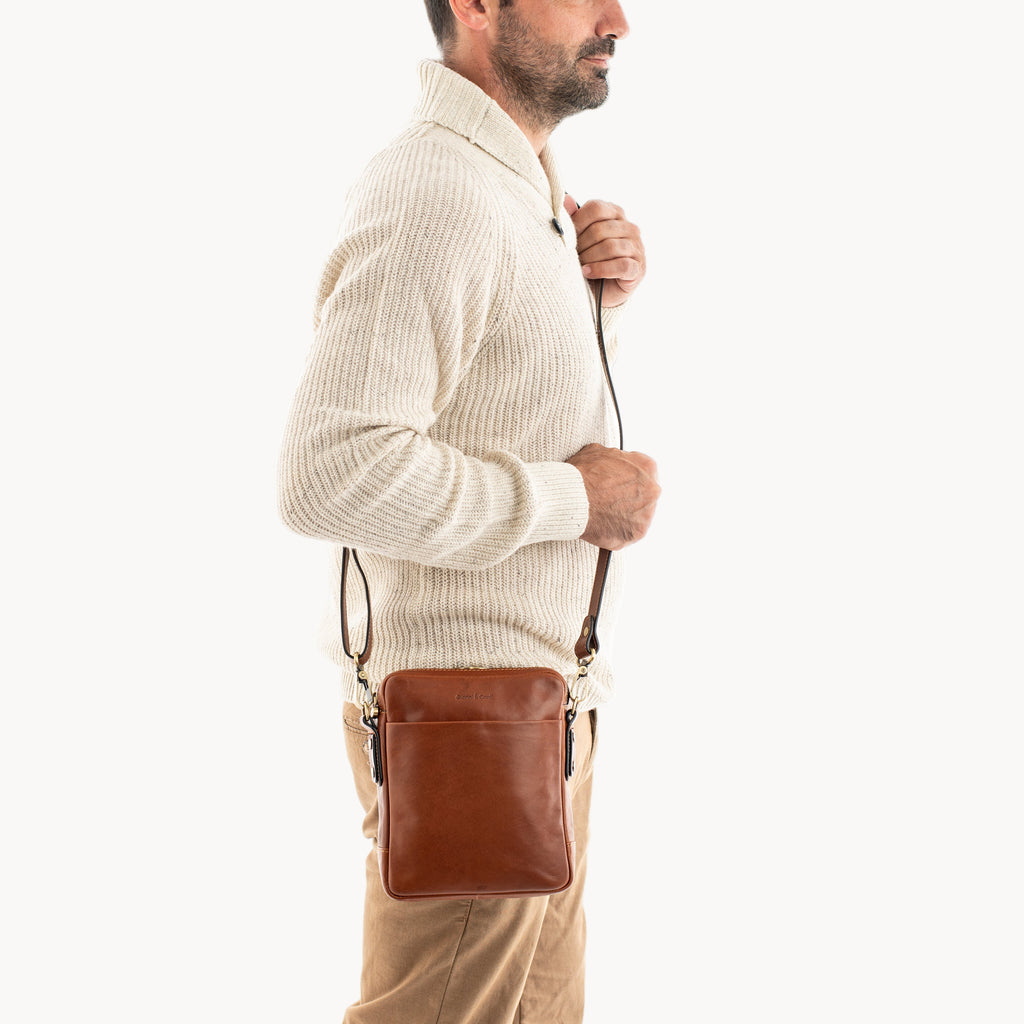 Man wearing a beige sweater and brown pants with a brown leather crossbody bag on a white background