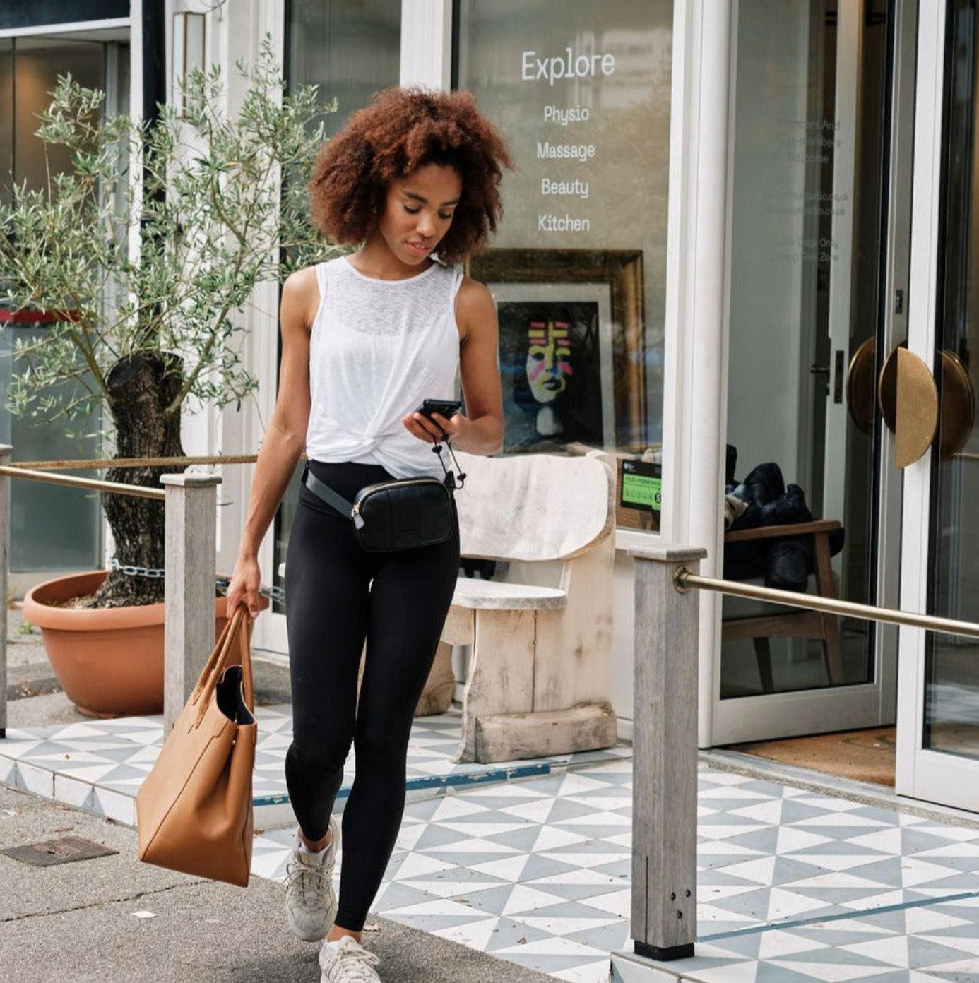 Woman walking outside a building, holding a phone and a brown handbag with a black belt bag around her waist.