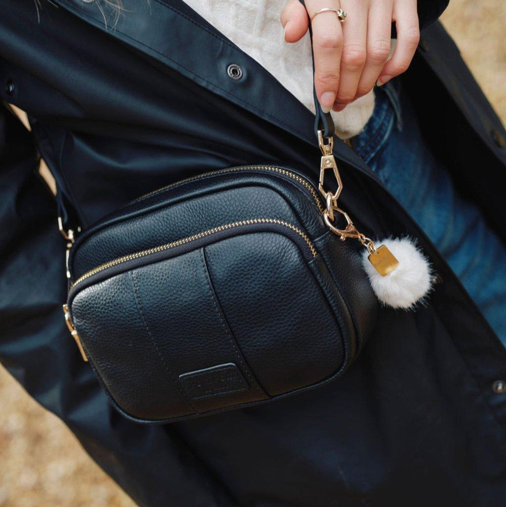 Black leather bag with a gold chain and white pom-pom charm held by a person wearing a dark jacket.