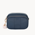 Navy blue crossbody bag with a gold zipper and white pom-pom charm on a white background