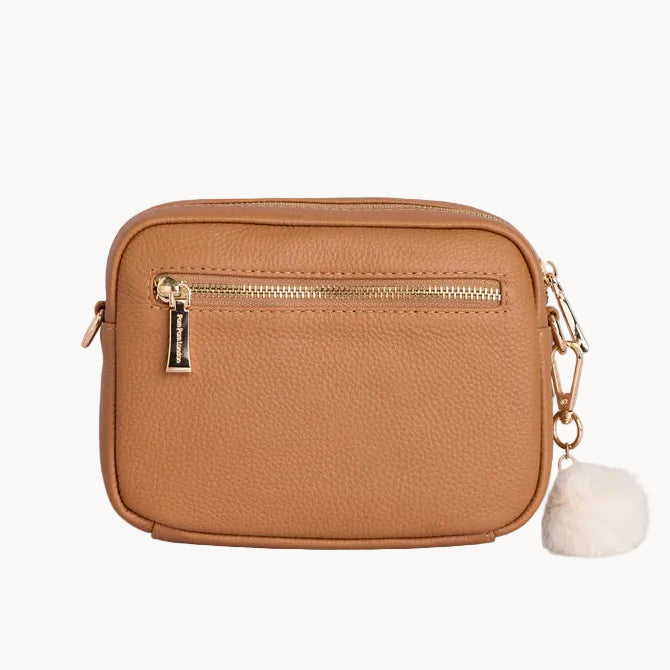 Tan leather crossbody bag with gold zipper and pom-pom on a light gray background
