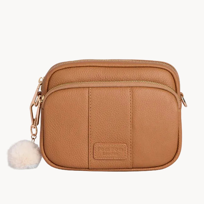 Tan leather crossbody bag with a pom-pom on a light gray background