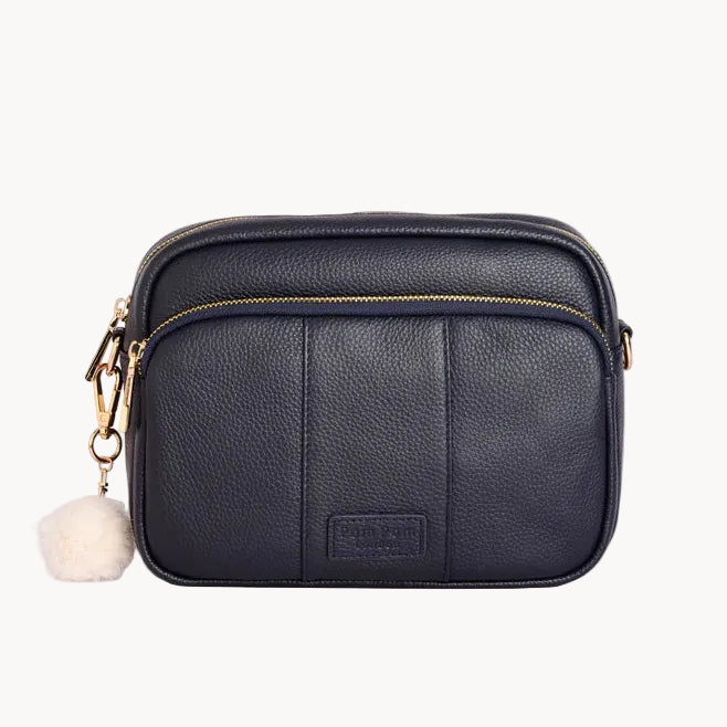 Navy leather crossbody bag with a pom-pom charm on a light gray background